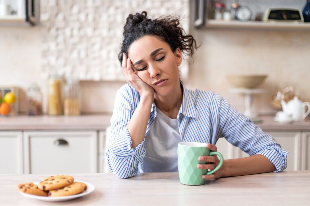 exhausted woman sitting at a kitchen table holding a mug reflecting on self-care mistakes that leave you exhausted