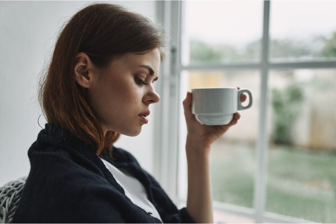 pensive tired woman sitting by window holding a warm cup wondering why self-care isn't working for you