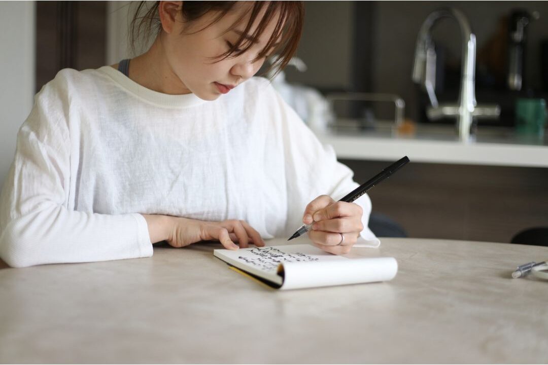 woman writing budget notes in a notebook at a sunny kitchen table making a plan to stop wasting money on these household items