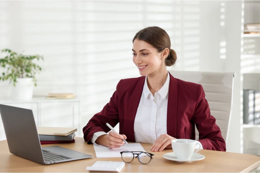 smiling woman sitting at a tidy desk writing in a budget planner to eliminate the hidden expenses quietly destroying your budget