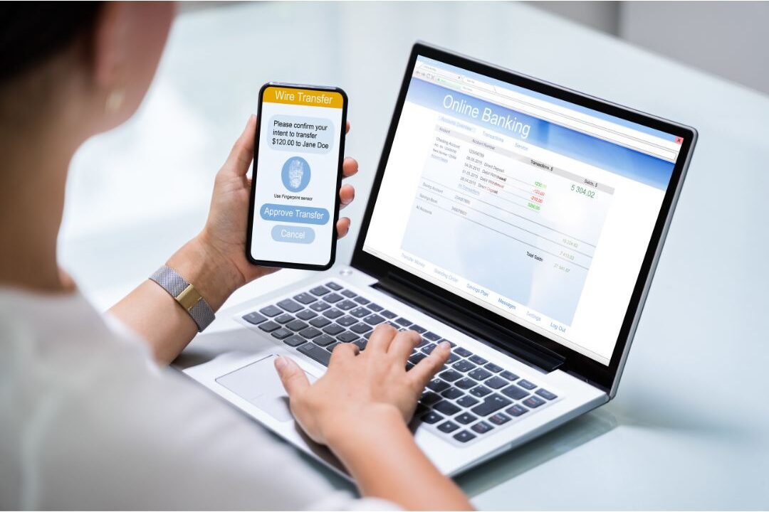 woman holding phone checking banking app near laptop setting up automated bill pay