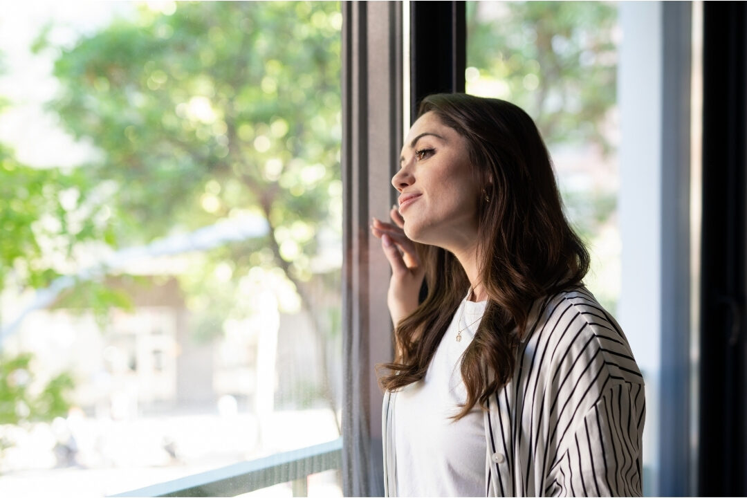 serene woman looking thoughtfully out sunlit window with notebook aligning work with natural energy peaks