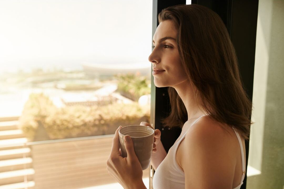 woman looking out sunny balcony window with coffee cup addressing maternal loneliness