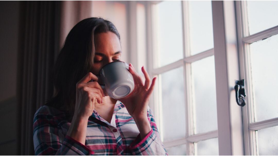 thoughtful woman looking out room window holding a ceramic mug reflecting on personal rest