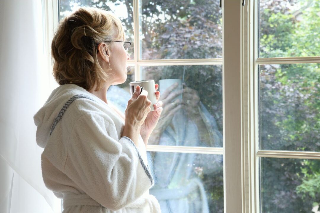 woman looking out balcony window with cup of tea in morning light reflecting on personal identity