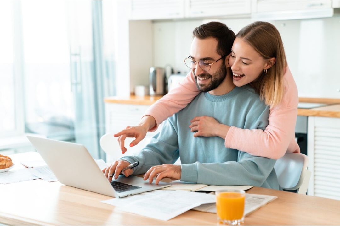 couple looking at laptop together at kitchen table smiling showing financial partnership