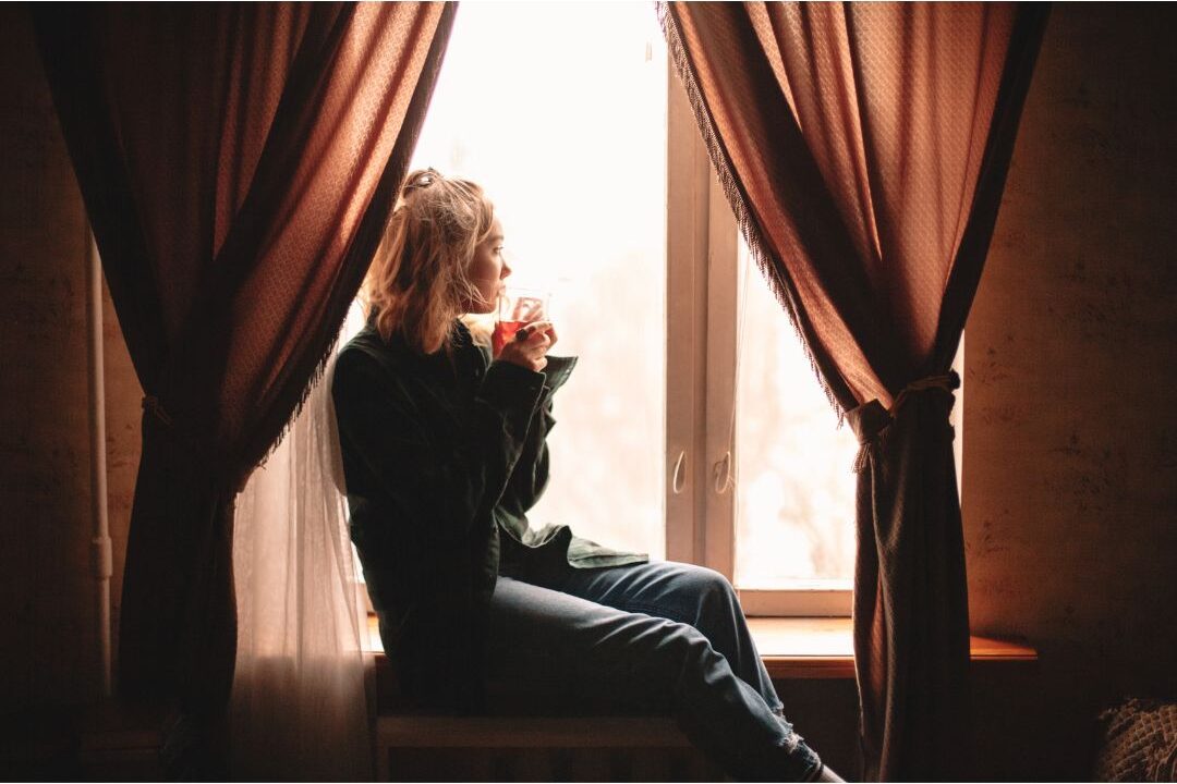 thoughtful woman sitting near sunny living room window with tea reflecting on the hard truth about being home all day
