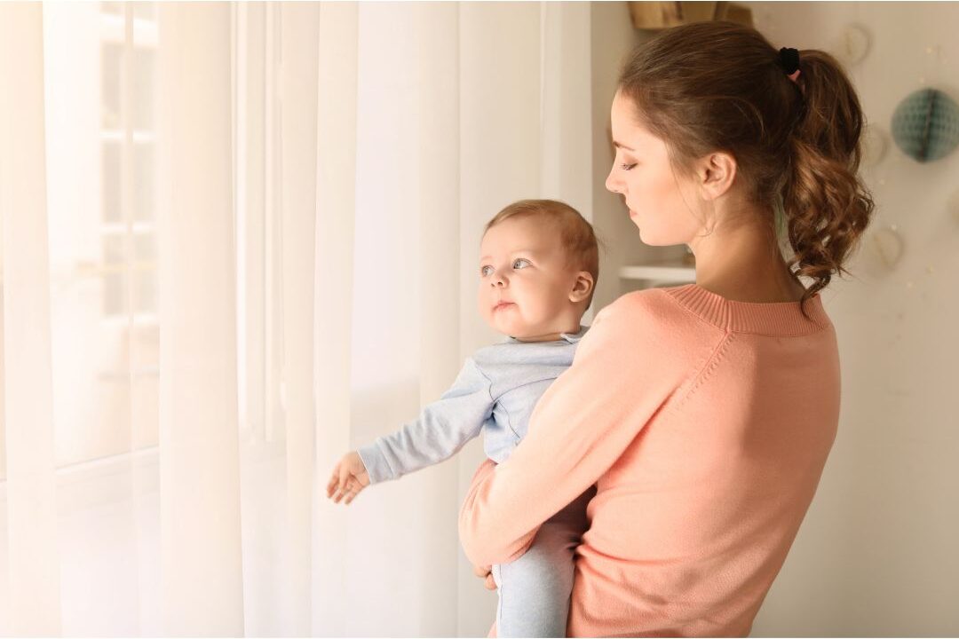 thoughtful mother holding baby near sunlit living room window learning about hidden challenges nobody warns moms about