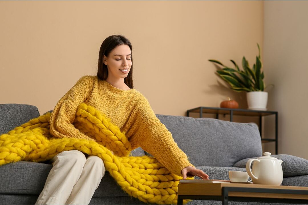 smiling woman sitting comfortably on couch wrapped in warm knit blanket enjoying restorative rest