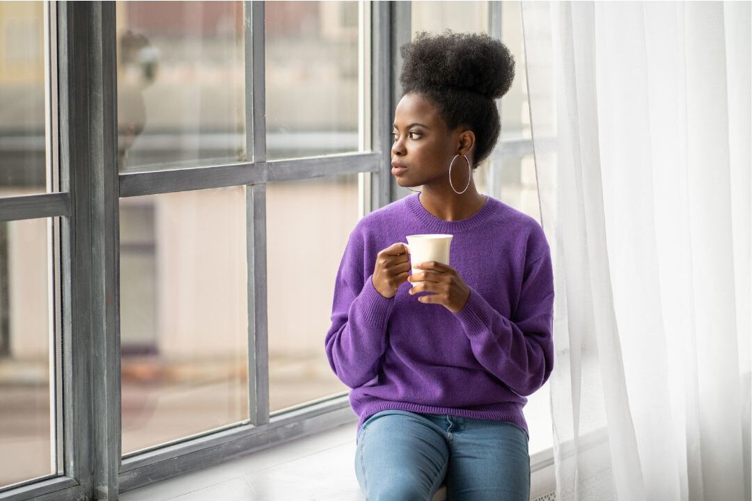 woman looking out a window thoughtfully while holding a coffee mug working through self-doubt