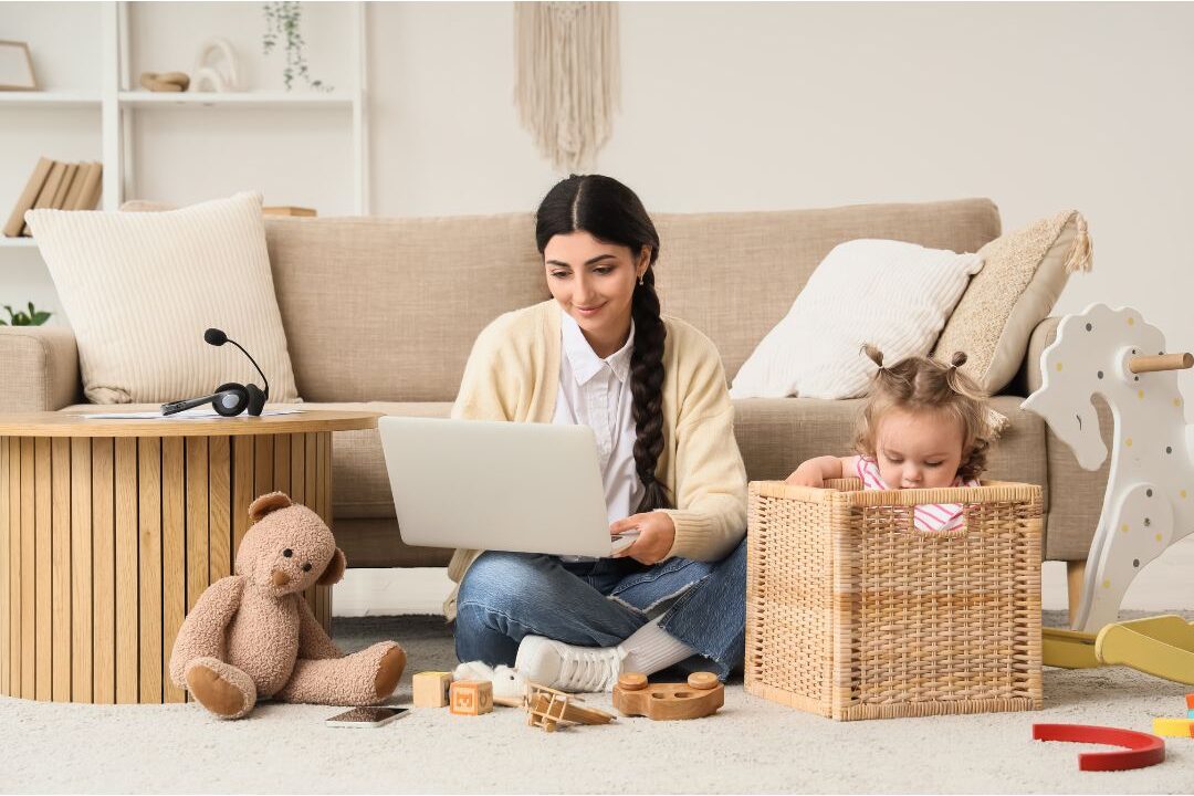 mother working on laptop at dining table with toddler toys nearby facing the hard truth about being home all day