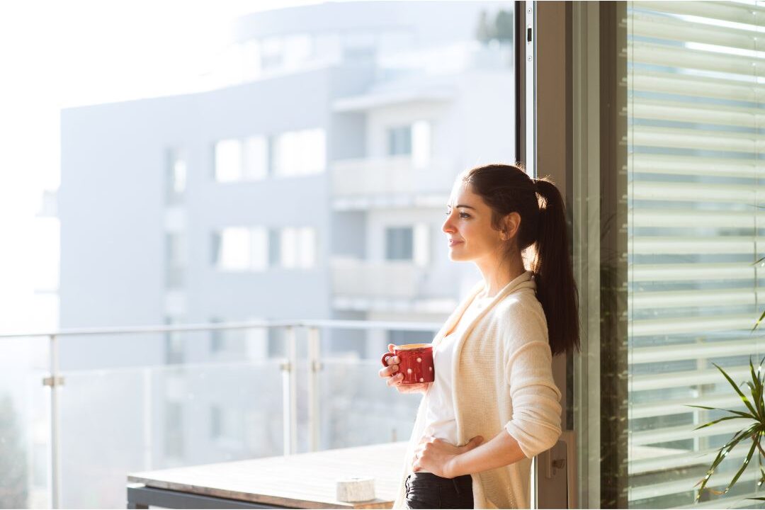 woman looking out sunny balcony window holding a warm mug reflecting on personal autonomy