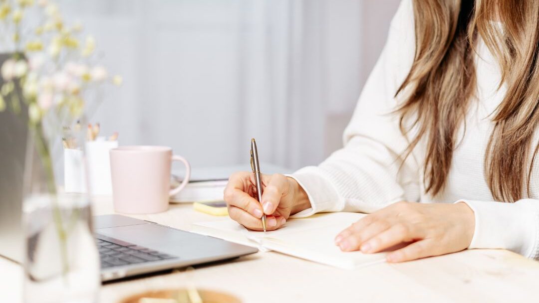 woman writing on a laptop with a notebook and coffee on her desk learning the biggest blogging mistakes beginners make