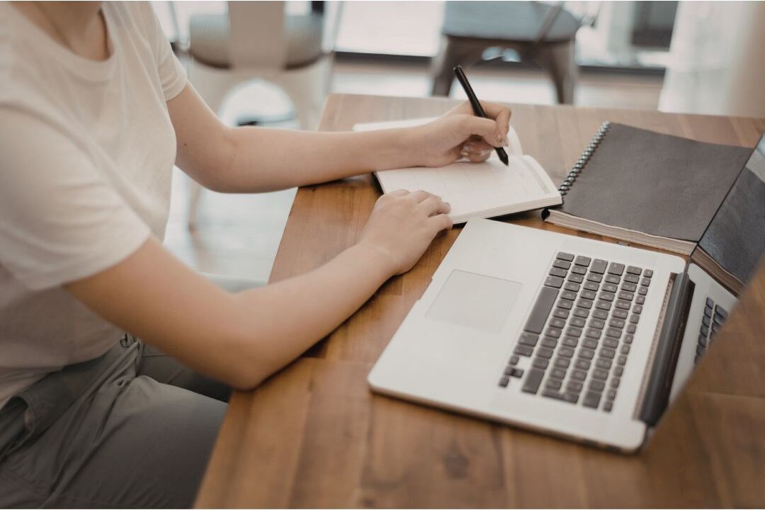 focused woman writing plans in a notebook next to a open laptop at a kitchen table