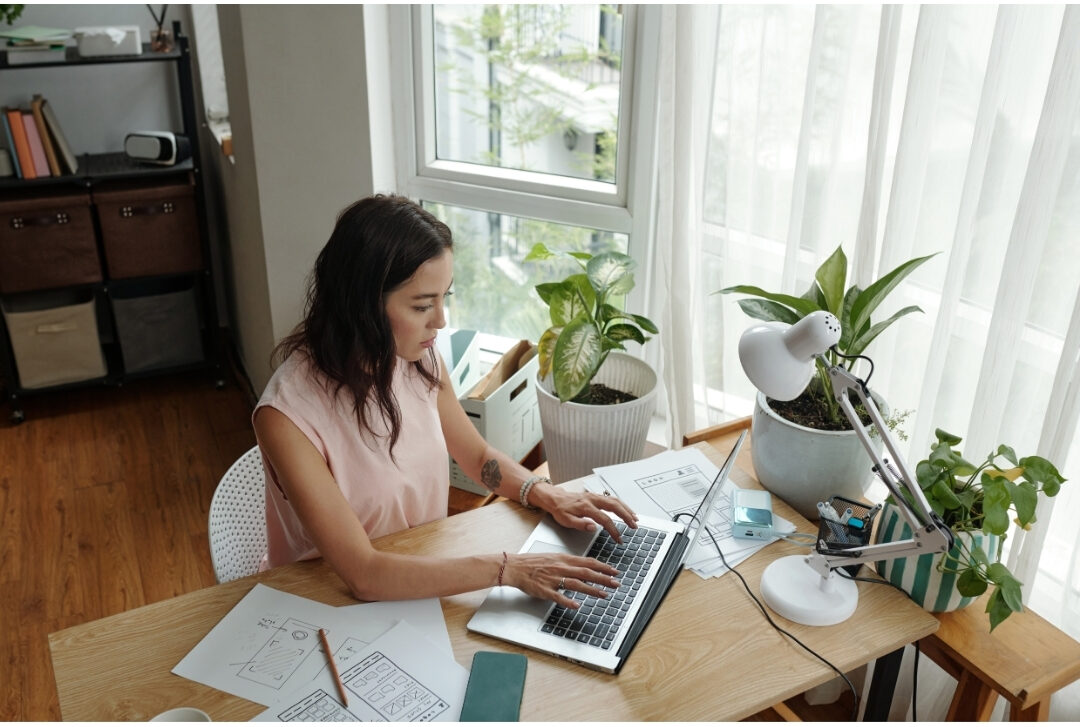 woman working on laptop at cozy dedicated wooden desk near window maintaining professional workspace