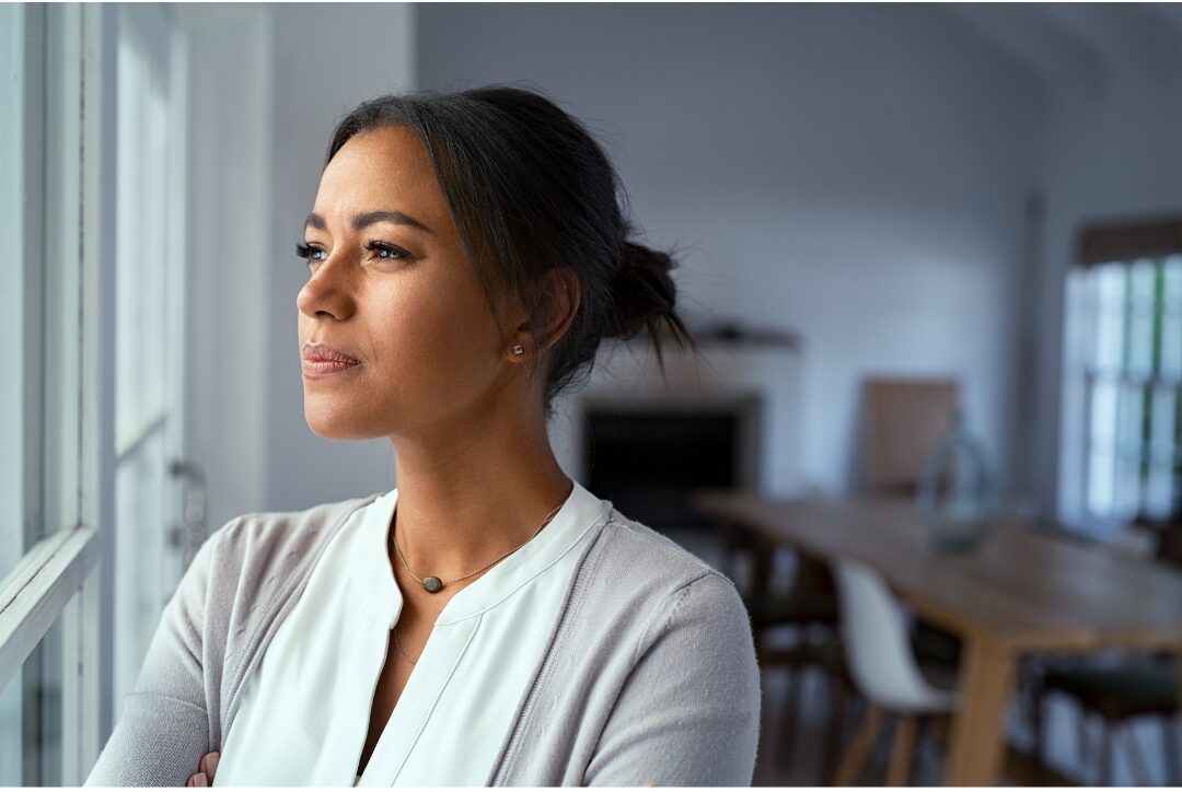 thoughtful woman standing near window looking outside with confident quiet expression identifying beliefs that keep moms playing small