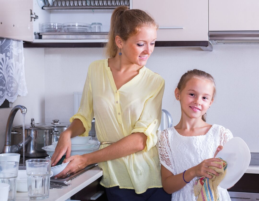 child putting away clean dishes in kitchen with mother smiling nearby delegating household tasks