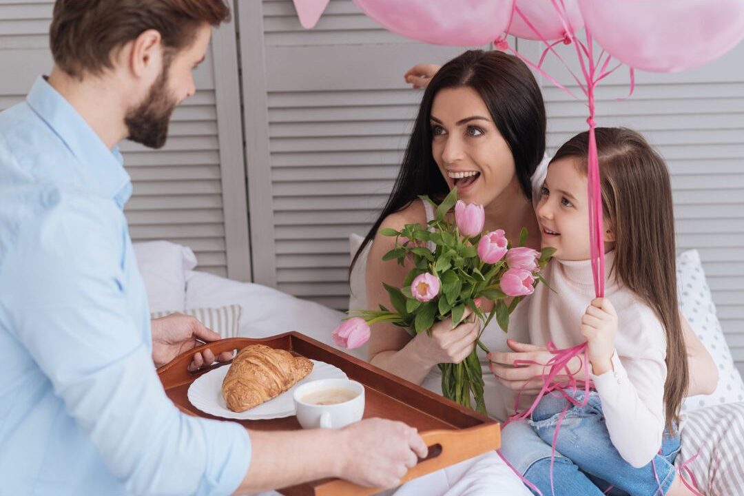 child sitting in bed holding breakfast tray with pancakes, candle, and balloon