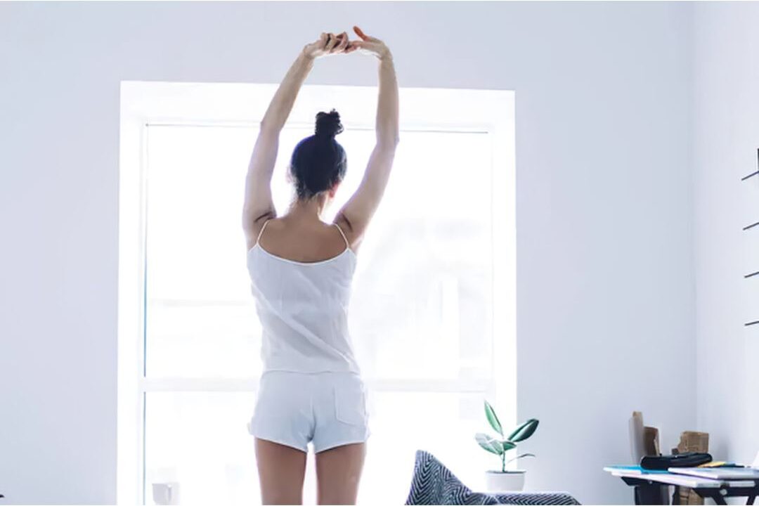 woman stretching arms overhead near sunny living room window displaying 45 daily habits that make life easier