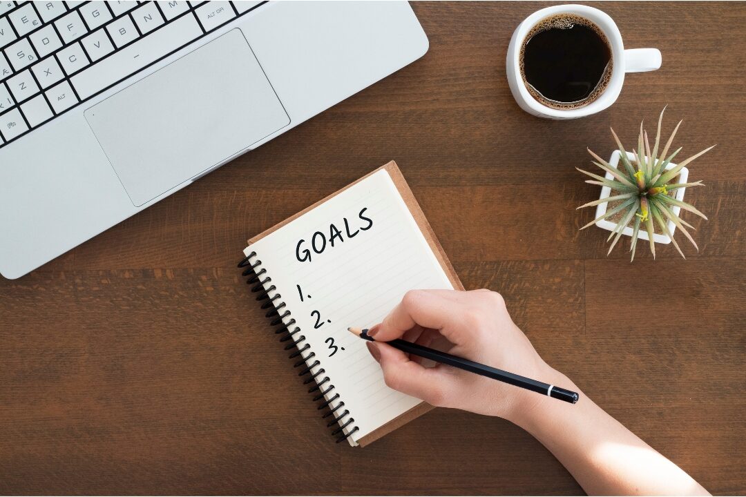 handwriting top three daily goals in paper notebook beside laptop defining daily targets