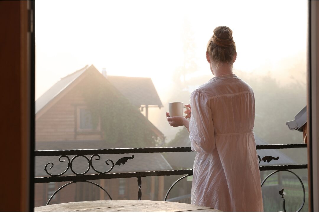 woman standing on sunlit balcony looking at sunrise with open arms stepping into full power