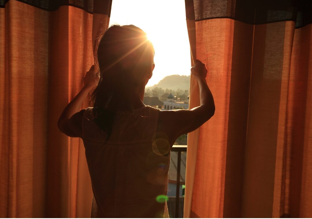 woman standing on sunlit balcony looking at sunrise with open arms knowing how to stop feeling like you're failing