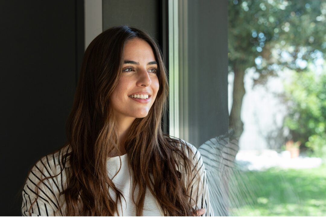 peaceful woman looking out sunlit window with serene smile understanding why comparison is stealing your joy